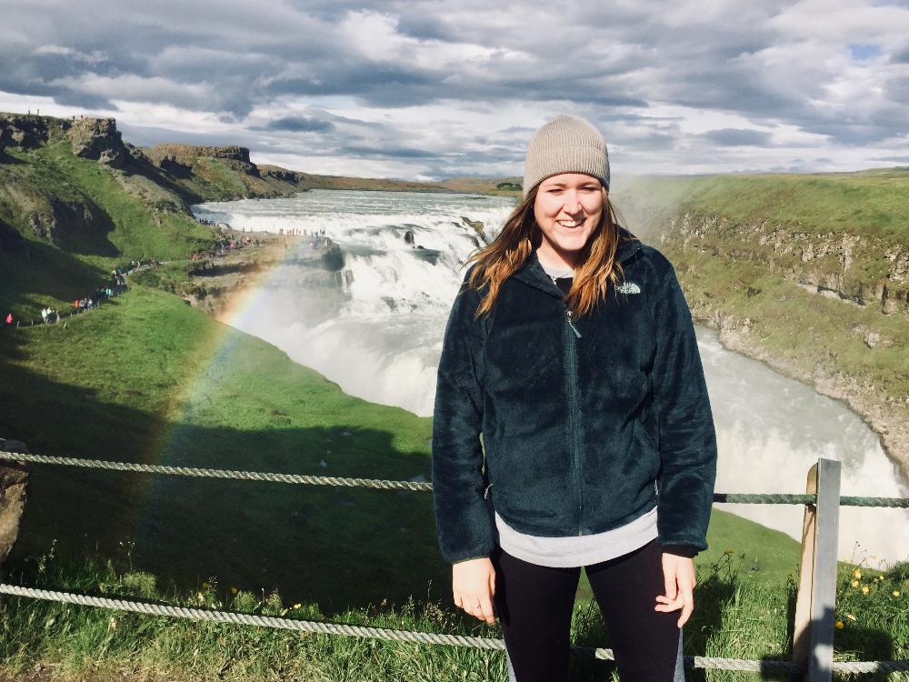 UTA student standing in front of river and waterfall with a rainbow