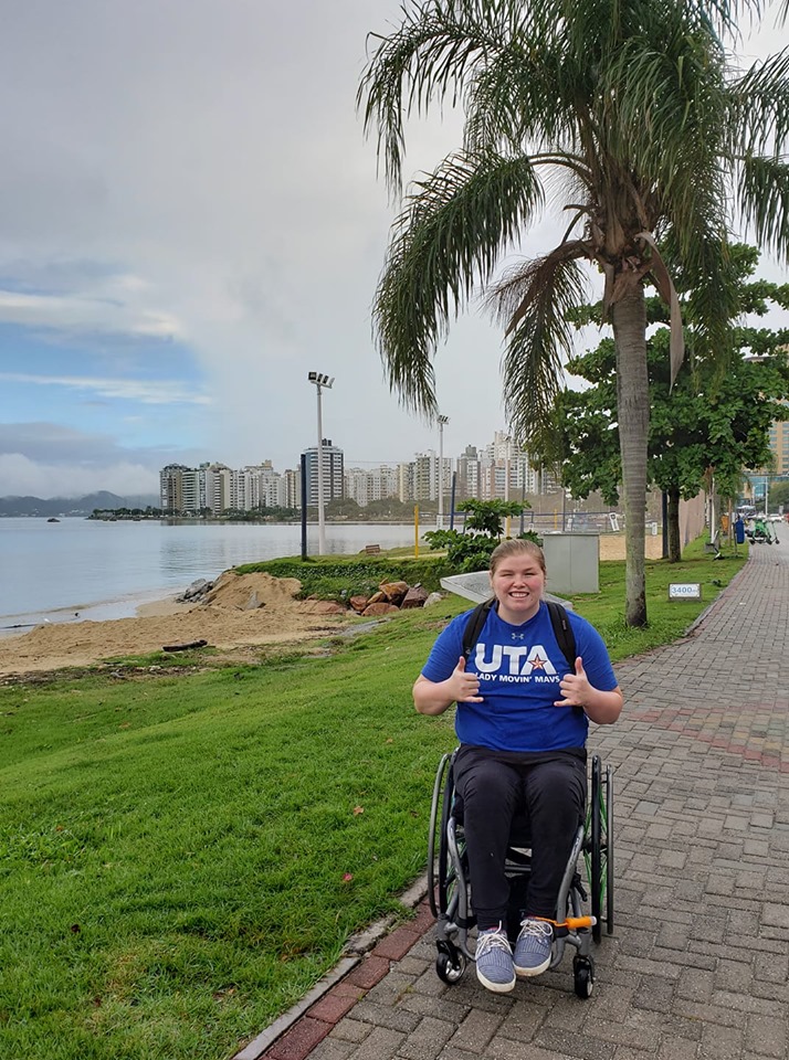 A student wearing a UTA t-shirt gives the "mav up" sign in front of a beach.